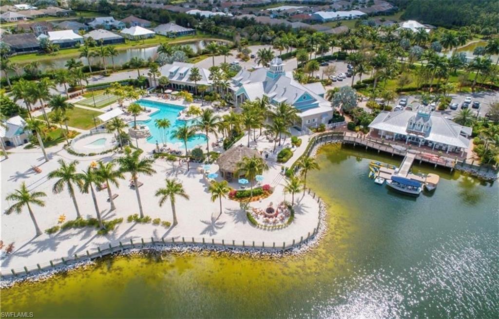 Community amenity center featuring a resort-style pool with a sun shelf, white sand beach, multiple buildings with gabled roofs, and a waterfront dock