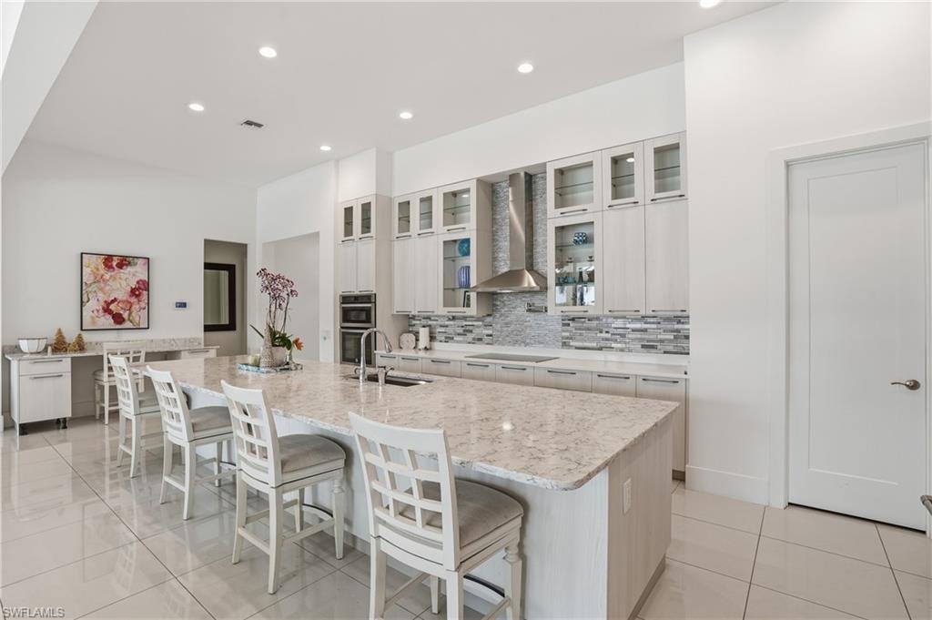 Spacious kitchen featuring a large island with a surface-mounted sink, built-in double ovens, a contemporary range hood, a tiled backsplash, and recessed lighting