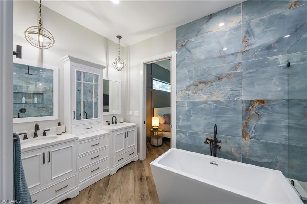 Bathroom featuring a freestanding soaking tub, large format blue tile accent wall, dual vanity with white cabinetry, wood-finish flooring, and black fixtures