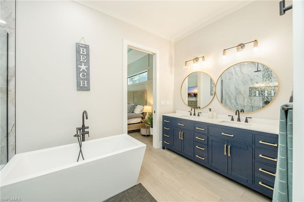 Spacious bathroom featuring a freestanding soaking tub with a floor-mounted faucet, a dual vanity with a dark blue cabinet base, gold hardware, and round mirrors, and wood-finish flooring