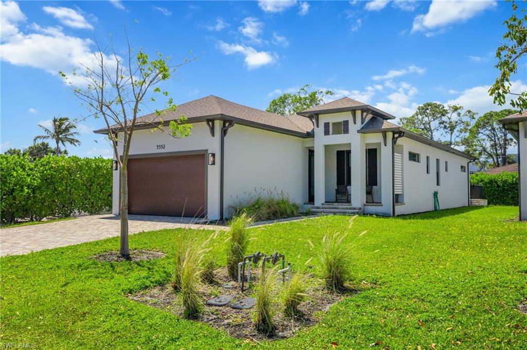 Contemporary stucco exterior with a prominent entry tower, dark trim accents, a paver driveway, an integrated garage with a wood-finish door, and manicured green landscaping