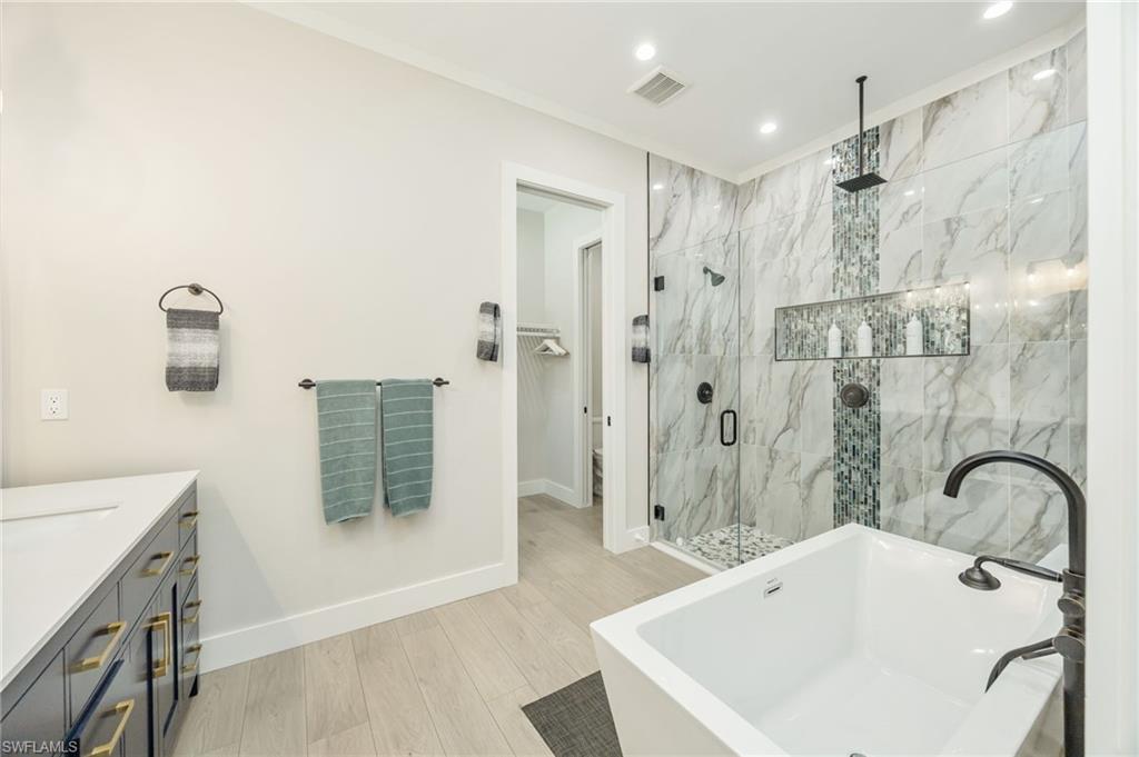 Contemporary bathroom featuring a freestanding soaking tub with a floor-mounted faucet, a spacious glass-enclosed shower with contrasting tile inlay and built-in niche, light-toned wood-finish flooring, and a dark vanity with a white countertop and gold-toned hardware