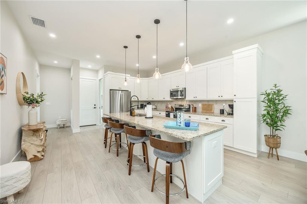 Modern kitchen featuring a large central island with seating, light granite countertops, white cabinetry with dark hardware, stainless steel appliances, and wood-finish flooring
