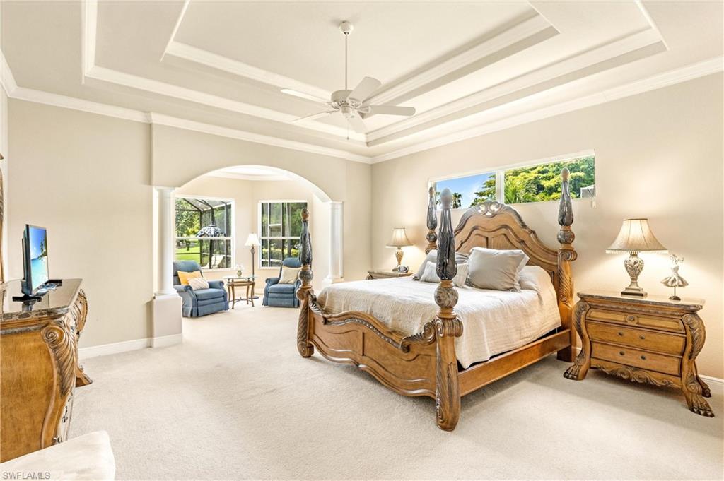 Spacious room with a tiered tray ceiling featuring a ceiling fan, light-colored carpeting, an arched entryway with columns, and multiple windows providing ambient light