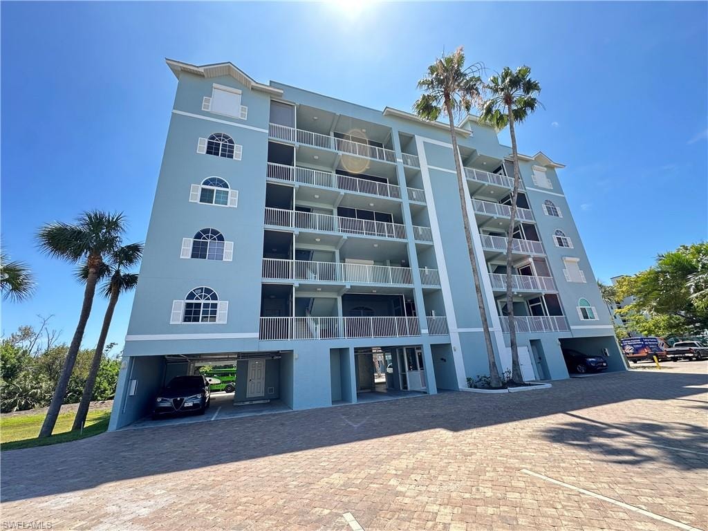 Multi-story residential building featuring a light blue exterior, multiple balconies, arched windows with white shutters, and ground-level covered parking