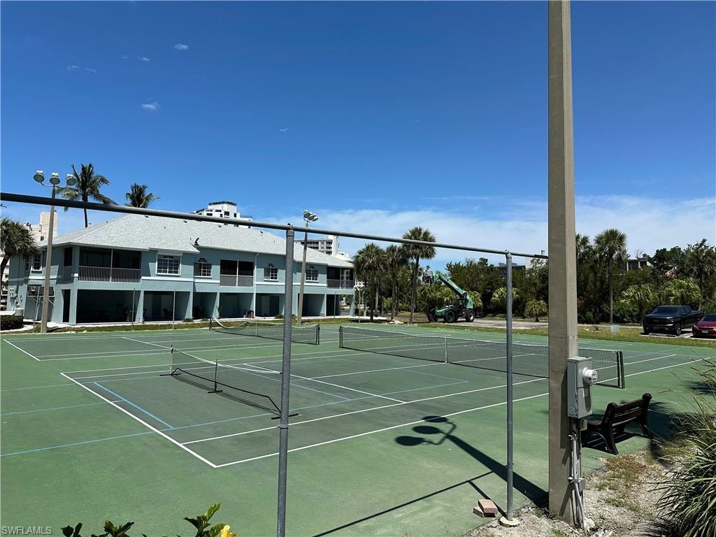 Tennis courts with perimeter fencing, a concrete light pole, and a distant two-story building featuring multiple balconies