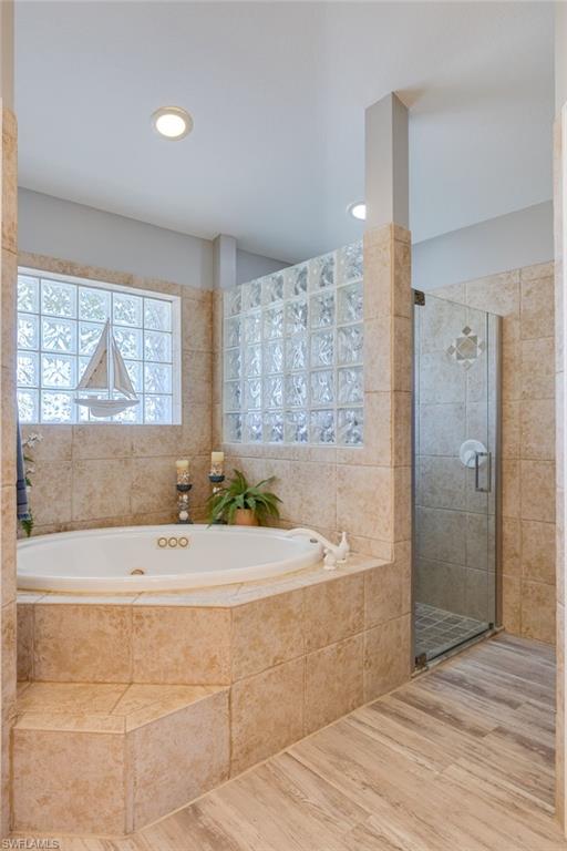 Soaking tub with a tiled surround, glass block window, glass-enclosed shower, wood-finish flooring, and recessed lighting