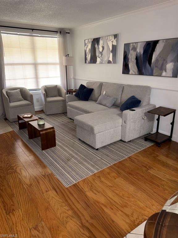 Living area featuring wood flooring, neutral wall tones, white crown molding, and a large window with blinds and sheer curtains