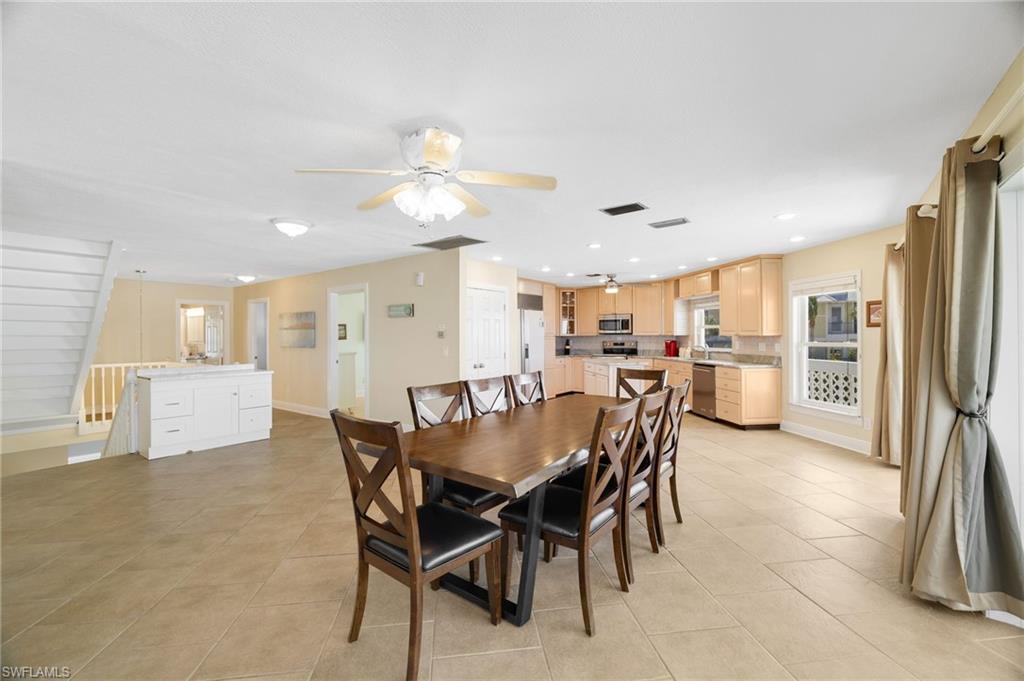 Open-concept interior featuring light-toned tile flooring, a staircase with white railing, and a kitchen with wood-finish cabinetry, stainless steel appliances, and a recessed lighting package - Virtually Edited Image