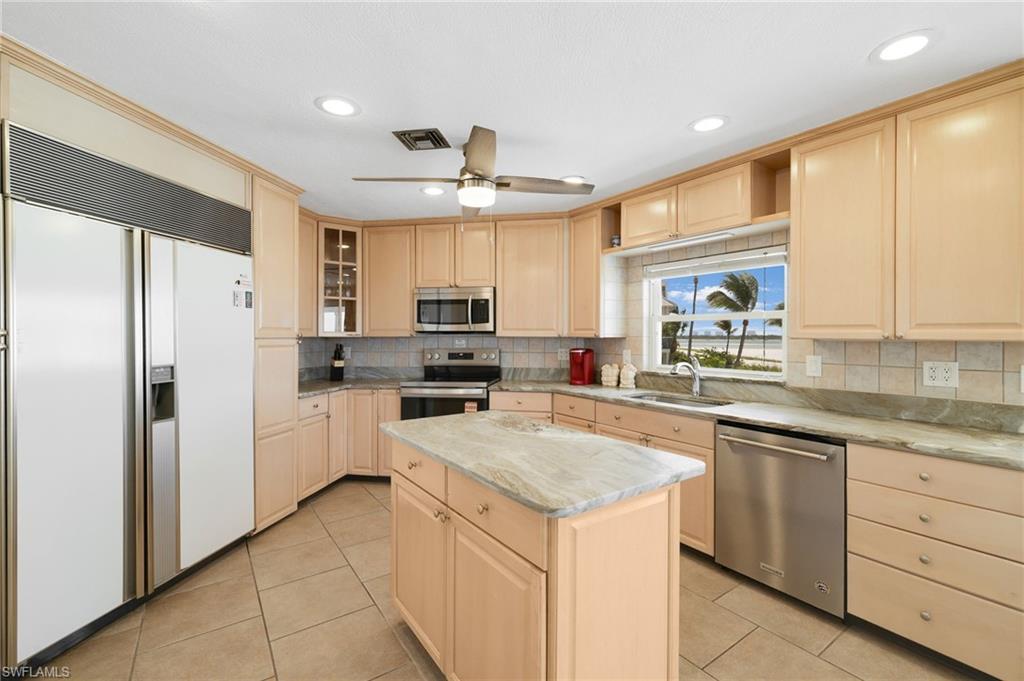 Spacious kitchen featuring a central island with a stone countertop, extensive cabinetry, and a window overlooking a beach with palm trees - Virtually Edited Image
