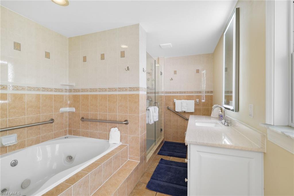 Bathroom featuring a jetted soaking tub, tiled shower with glass enclosure, single vanity with light-colored countertop, and a back-lit mirror - Virtually Edited Image