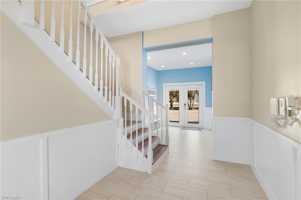 Entryway featuring a white banister staircase with wood-finish treads, a white wainscoting wall, and light-toned tile flooring - Virtually Edited Image