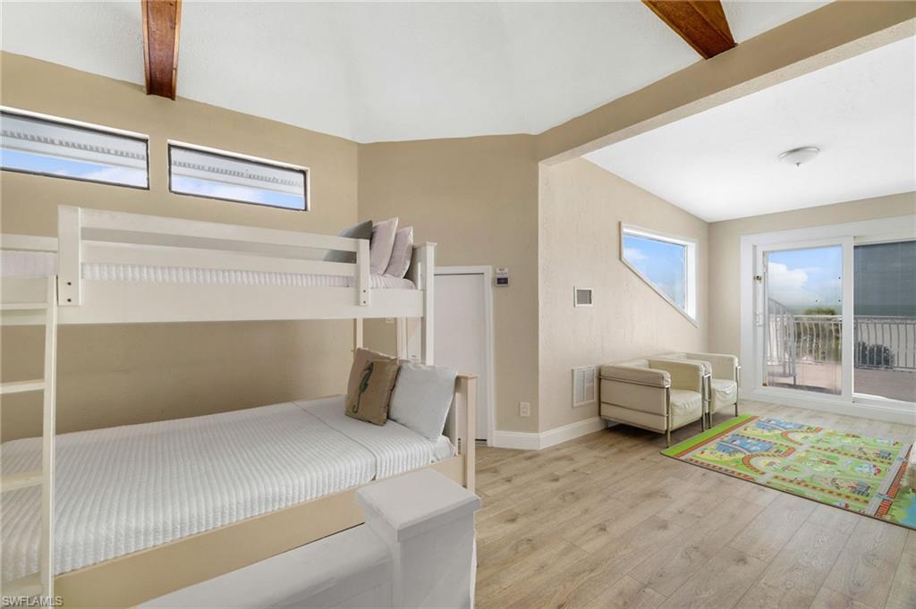 Spacious room featuring wood-finish flooring, a vaulted ceiling with exposed wood beams, and a sliding glass door opening to an outdoor balcony with a metal railing - Virtually Edited Image