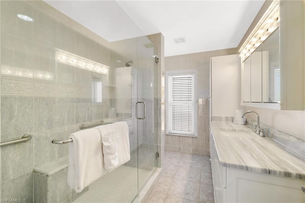 Spacious bathroom featuring a glass-enclosed shower with integrated bench seating, a dual vanity with a patterned countertop, contemporary light fixtures, and tiled walls and flooring - Virtually Edited Image