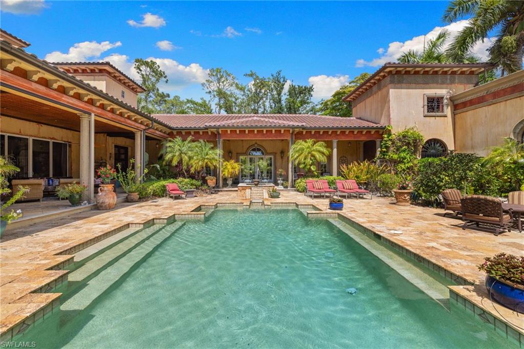 Spacious outdoor living area featuring a rectangular pool with integrated steps, extensive stone patio, covered loggia with columns, and lush landscaping