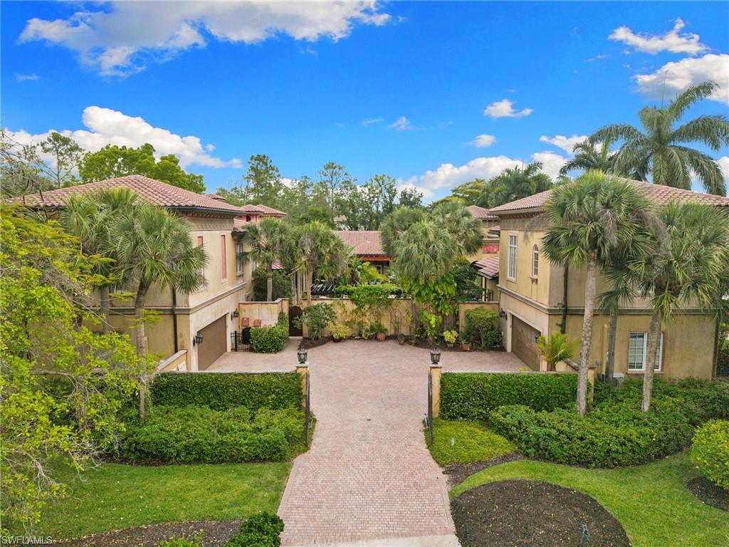 Mediterranean-style residence featuring a patterned paver driveway, terracotta roof tiles, stucco exterior, integrated garage doors, and lush tropical landscaping
