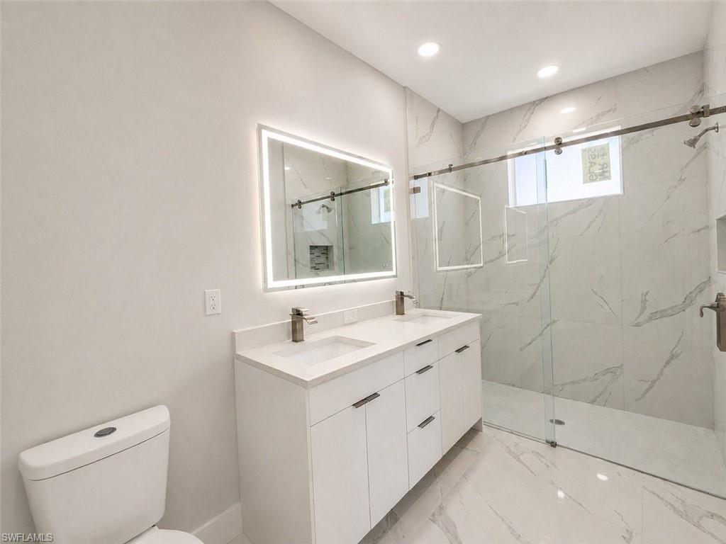 Bathroom featuring a dual vanity with white cabinetry, a backlit mirror, a glass-enclosed shower with a rainfall showerhead, and large format white tile flooring and walls