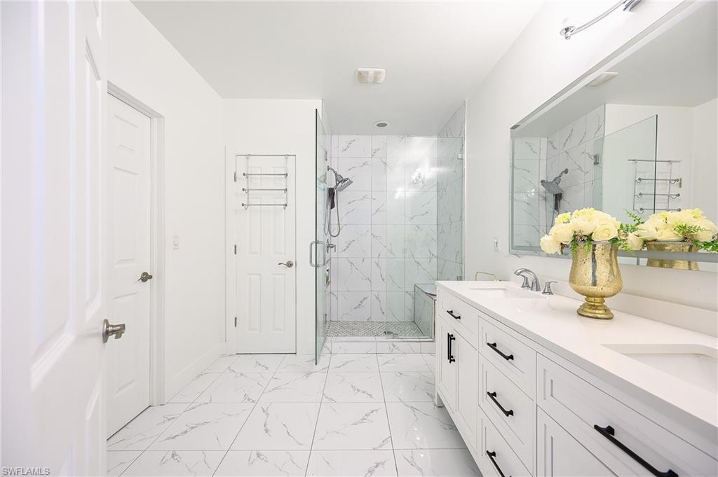 Spacious bathroom featuring a double vanity with ample storage, a large mirror, a glass-enclosed shower with a built-in bench, and white tile flooring with veining