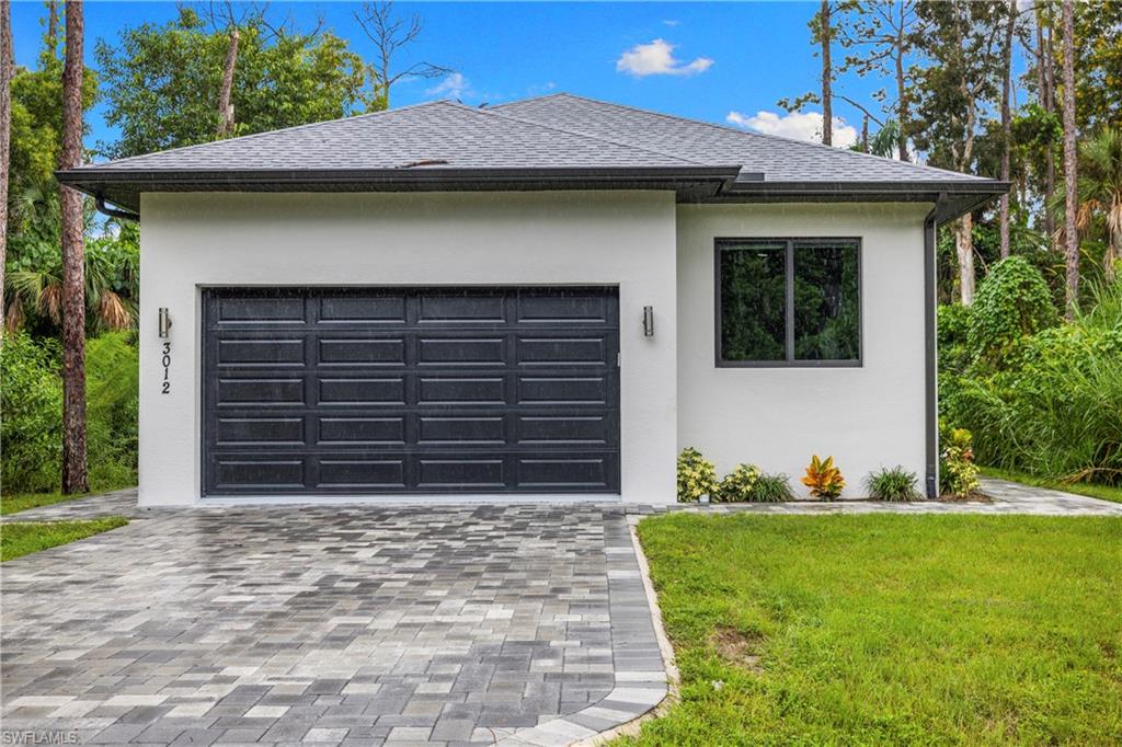Contemporary facade featuring a dark gray paneled garage door, white stucco exterior, black-framed window, and a paver driveway