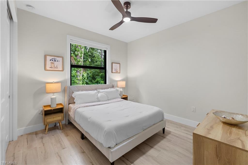 Bedroom featuring light-toned wood-finish flooring, a contemporary ceiling fan, and a window framing lush greenery