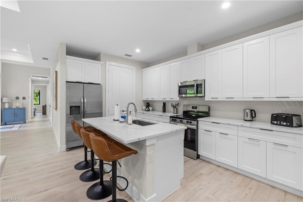Kitchen featuring an island with a built-in sink, stainless steel appliances, white cabinetry, and light wood-finish flooring