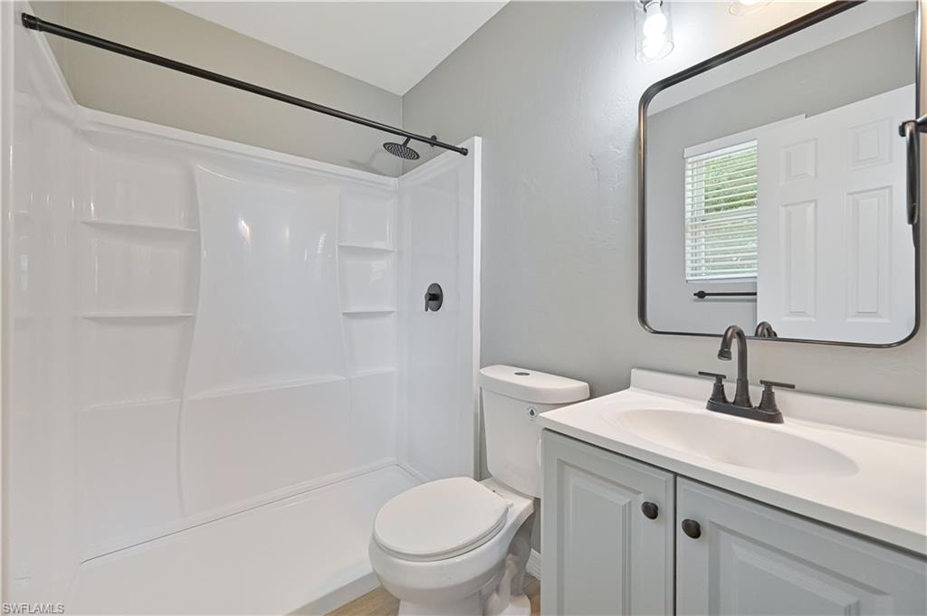 Bathroom featuring a white shower surround with built-in shelving, a single vanity with light gray cabinetry and dark hardware, a rectangular mirror with a dark frame, and wood-finish flooring