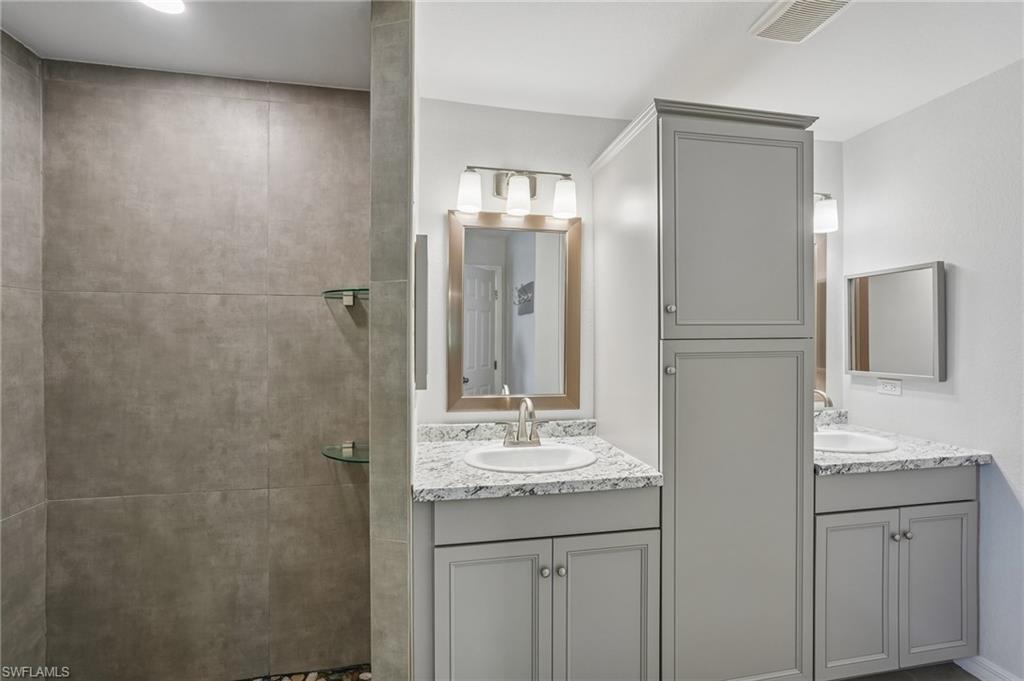Bathroom featuring a walk-in shower with large format tile, dual vanity sinks with light-colored countertops, grey cabinetry, and overhead lighting fixtures