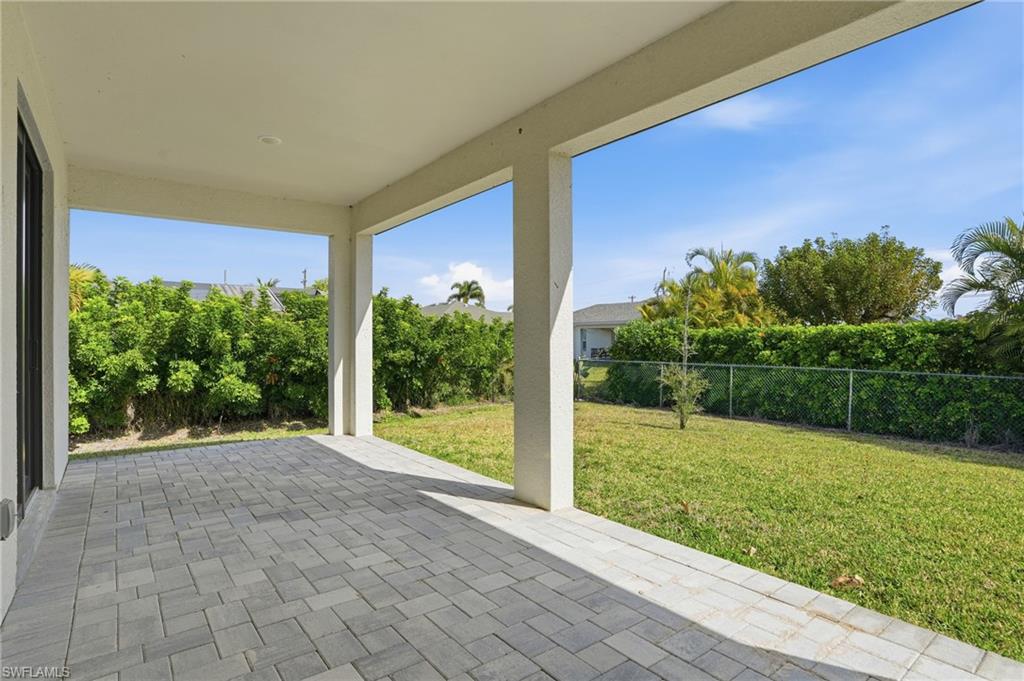 Covered patio featuring an expansive paver surface, integrated lighting, and direct access to a grassy yard with established landscaping and a chain-link fence
