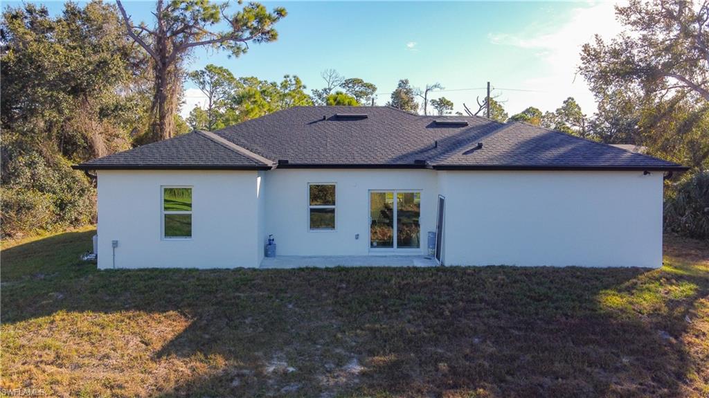 Rear exterior featuring a white stucco finish, a dark shingle roof, multiple windows, a sliding glass door, and a concrete patio