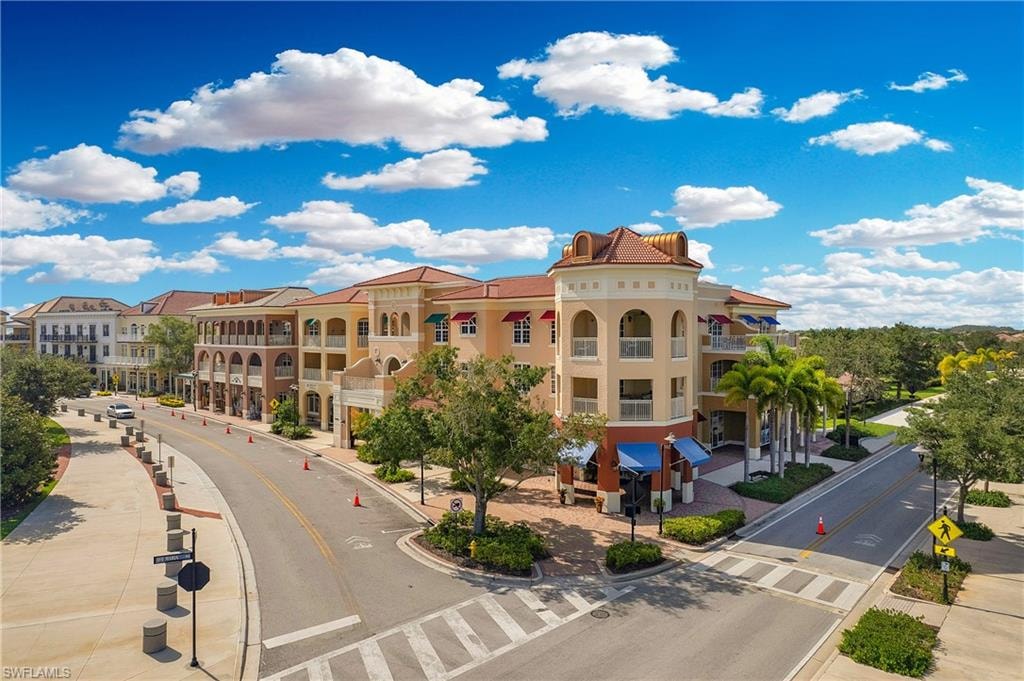 Multi-story building featuring Mediterranean-inspired architecture, prominent corner turret, individual balconies, ground-level retail storefronts with awnings, and surrounding mature landscaping
