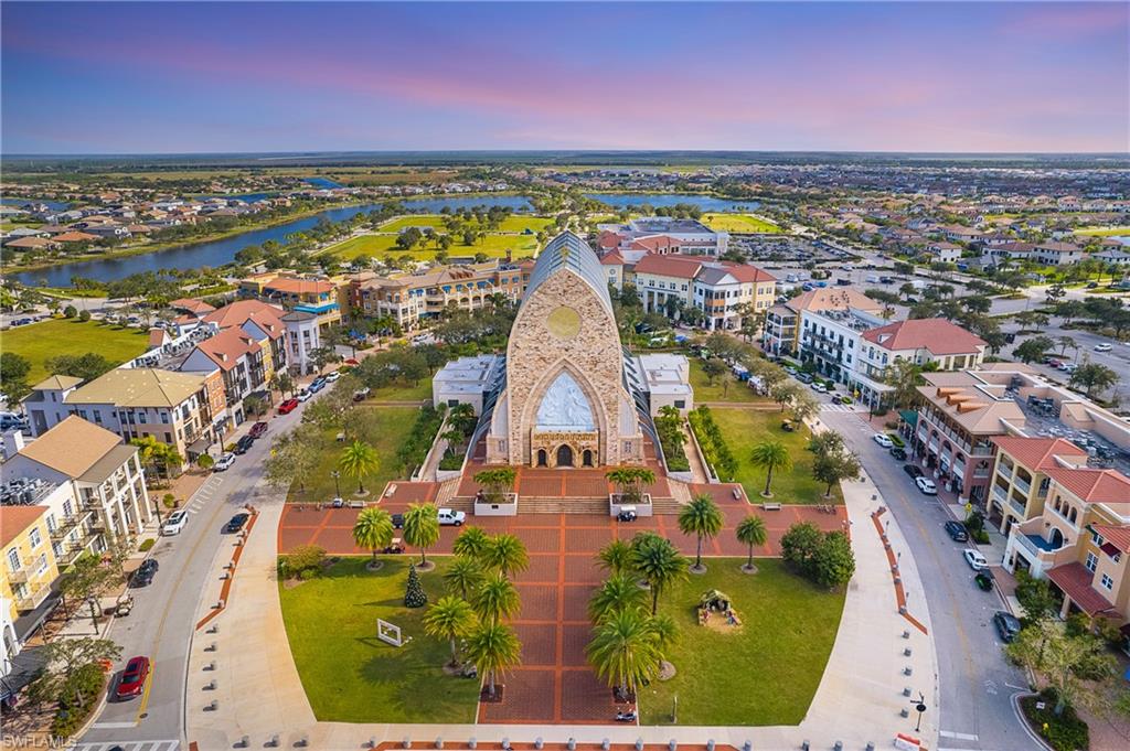 Expansive town center featuring a prominent architectural structure with a unique facade, surrounded by multi-story buildings with varied architectural styles, manicured green spaces, mature palm trees, and brick-patterned hardscaping
