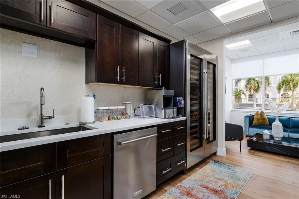 Kitchenette featuring dark wood-finish cabinetry, a stainless steel sink with a gooseneck faucet, light-toned countertops, and a built-in stainless steel dishwasher