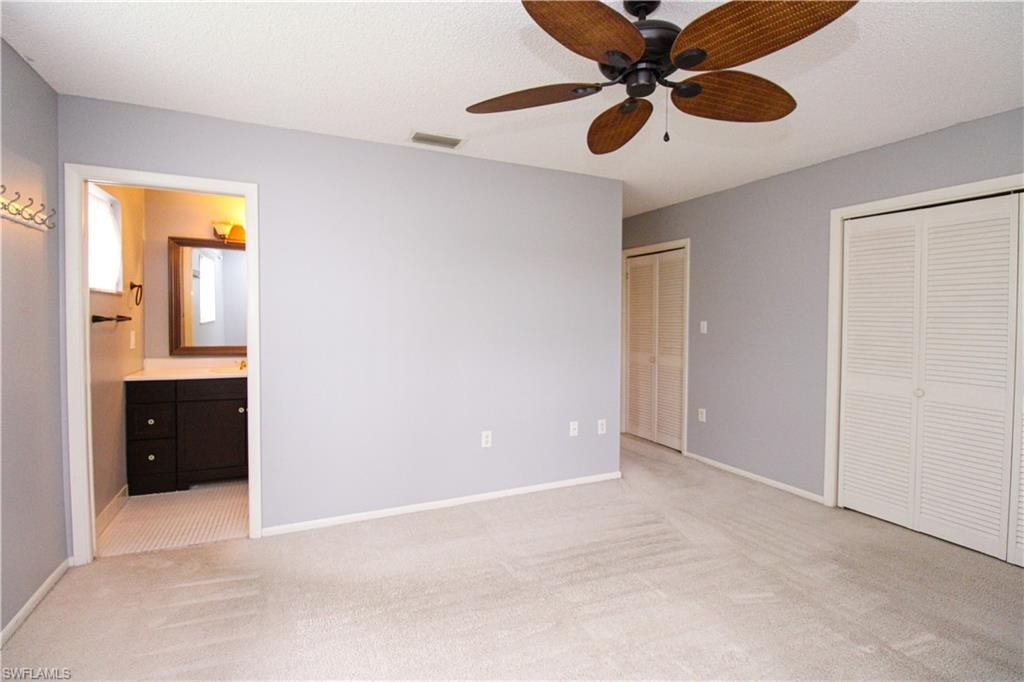 Unfurnished bedroom with two closets, light colored carpet, ensuite bath, and ceiling fan