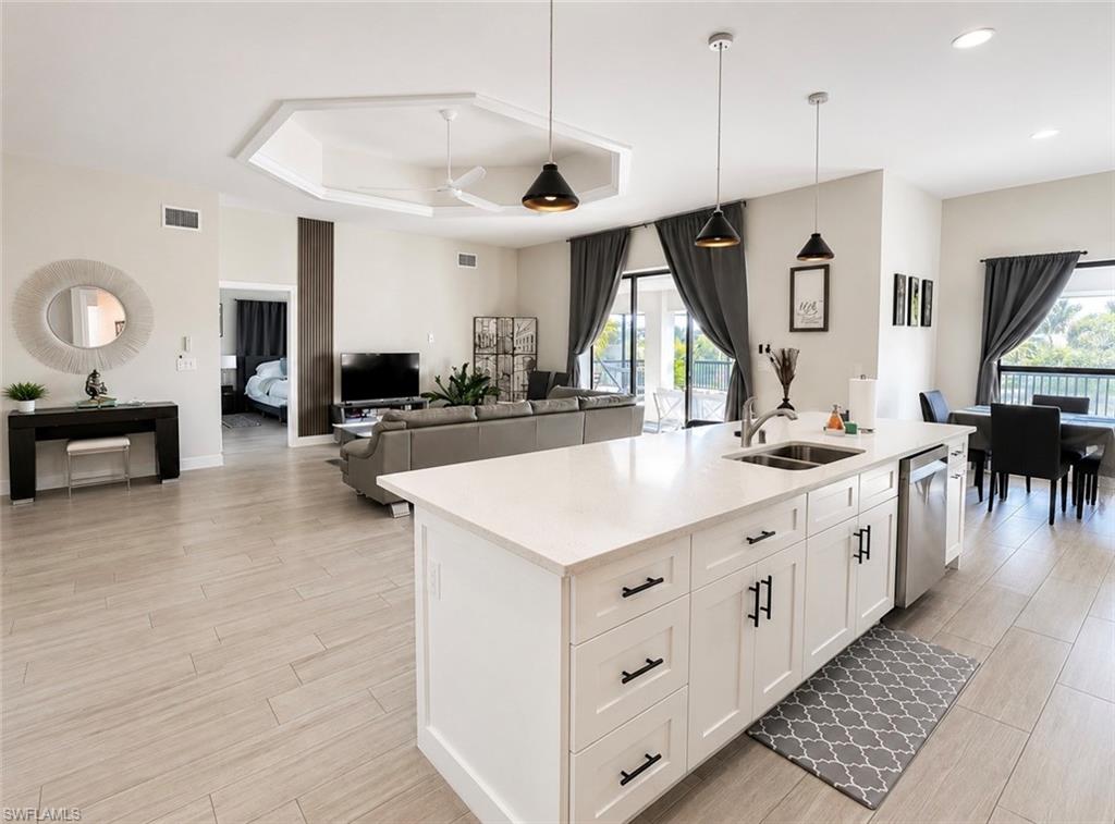Open-concept living space with light-toned wood-finish flooring, white kitchen island with undermount sink, stainless steel dishwasher, and pendant lighting