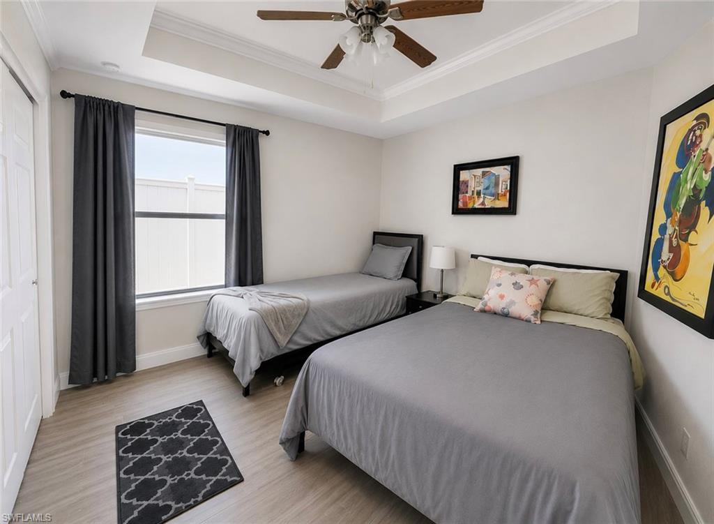 Secondary bedroom featuring two twin beds with gray upholstered frames, wood-finish flooring, a ceiling fan with integrated lighting, large window with dark curtains providing natural light, and framed artwork on neutral walls