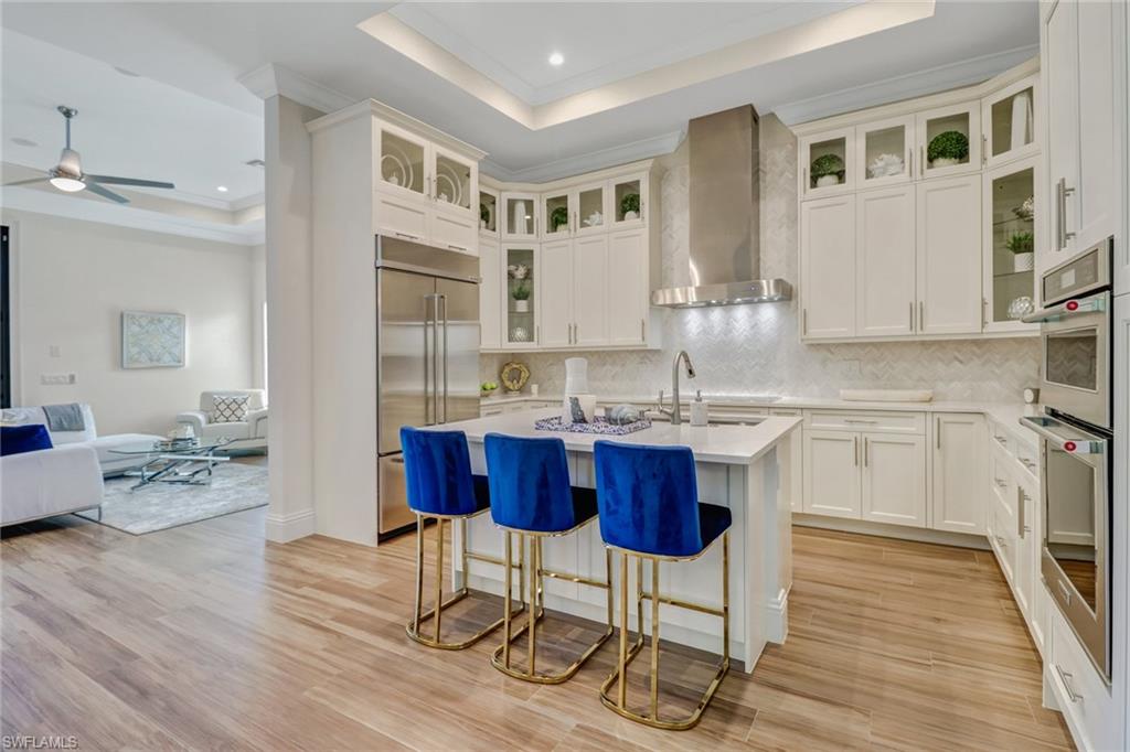 Kitchen with sink, wall chimney exhaust hood, an island with sink, a tray ceiling, and stainless steel built in refrigerator