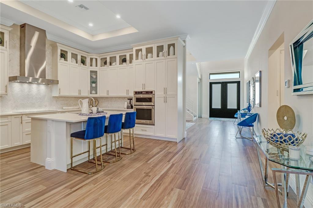 Kitchen with a raised ceiling, wall chimney range hood, an island with sink, double oven, and a breakfast bar area