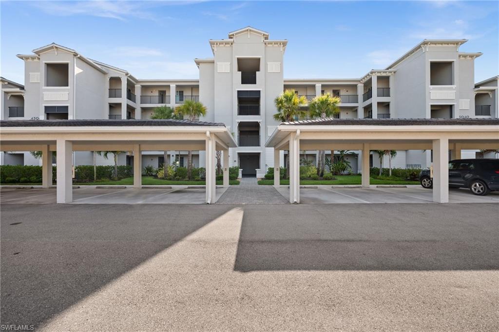 Multi-story building featuring balconies with dark railings, a central entrance with a paved walkway, and landscaping with palm trees