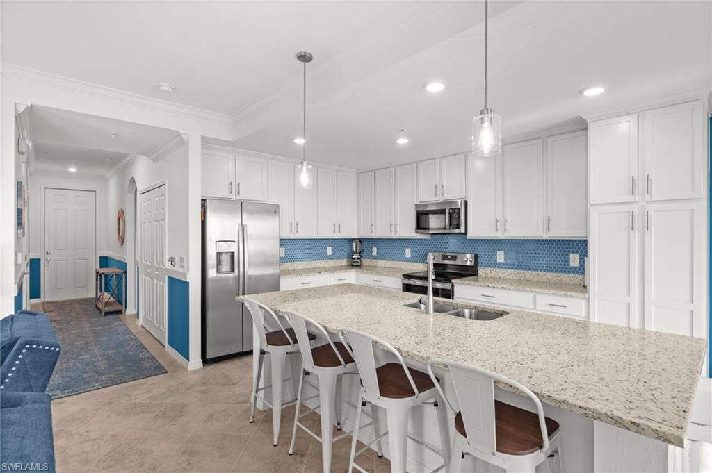 Spacious kitchen featuring white cabinetry, stainless steel appliances, light-toned countertops, a blue mosaic tile backsplash, and an expansive island with seating