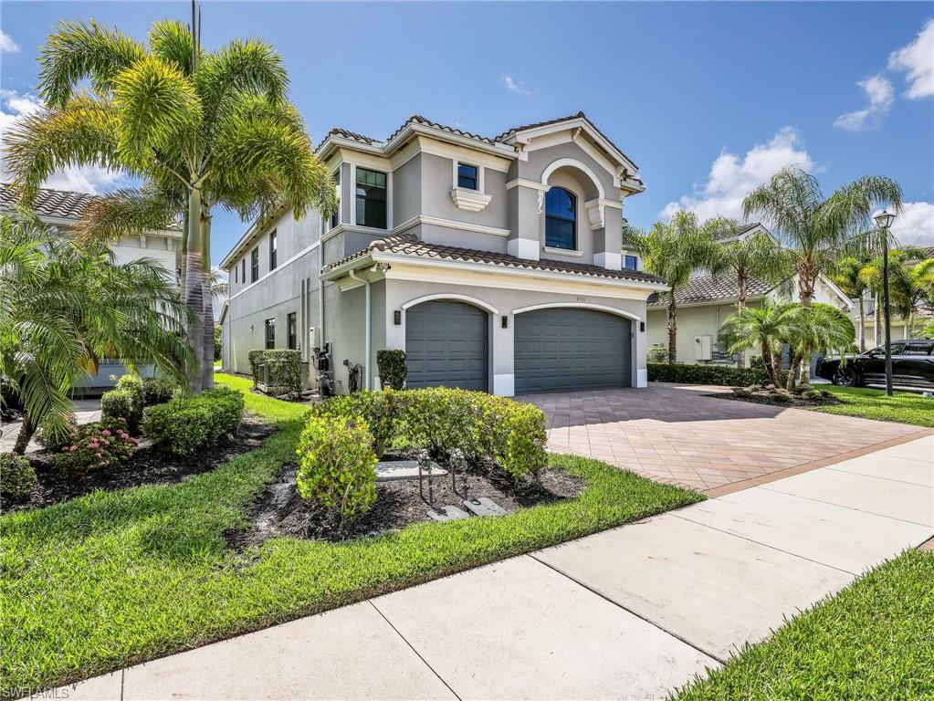 Two-story stucco exterior with arched window accents, tile roof, dual garage doors, paver driveway, and mature landscaping