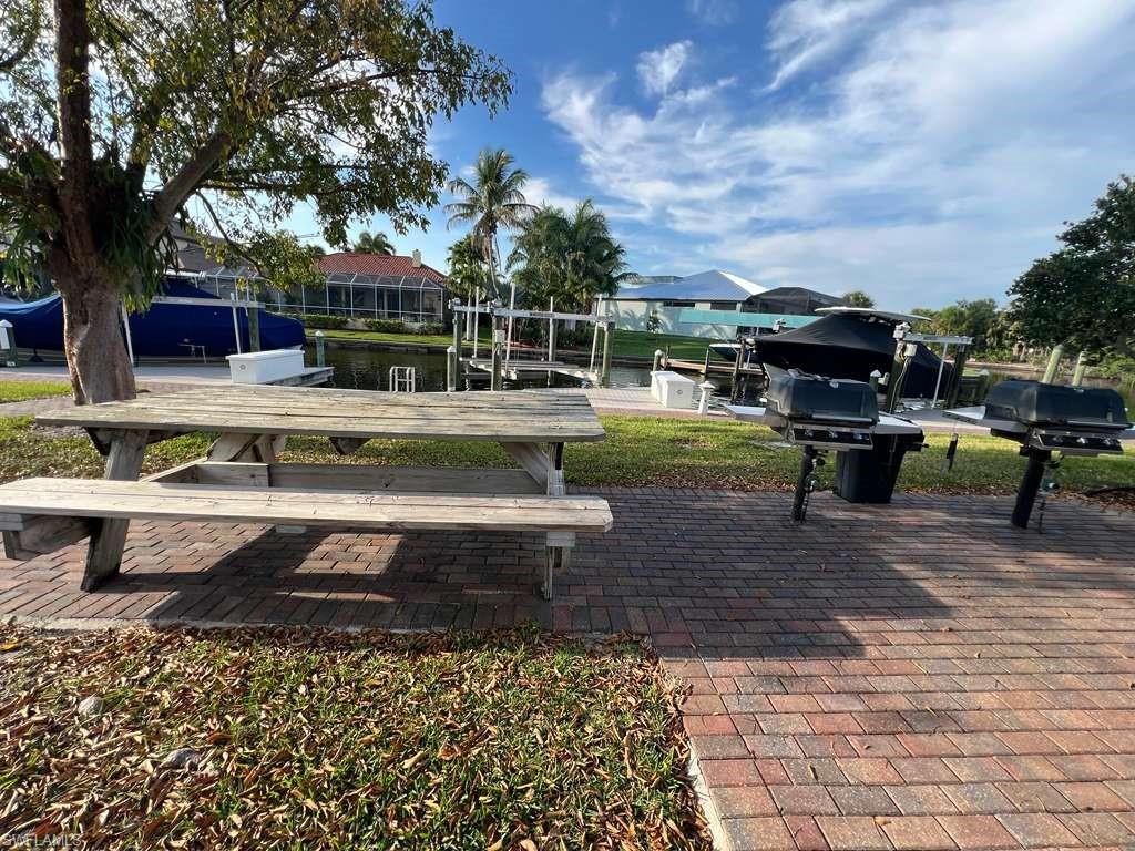 Paved brick patio with picnic table and dual barbecue grills, set against a canal with boat docks and tropical landscaping