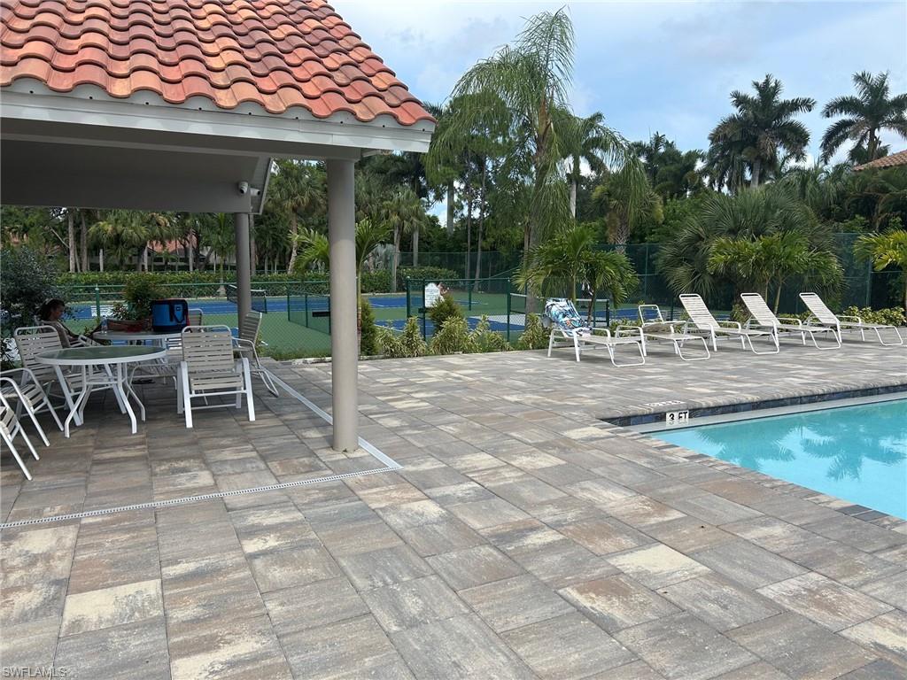 Poolside patio featuring multi-toned paver flooring, a covered seating area with a terracotta-style tiled roof, a swimming pool with a dark coping, and a tennis court visible in the background