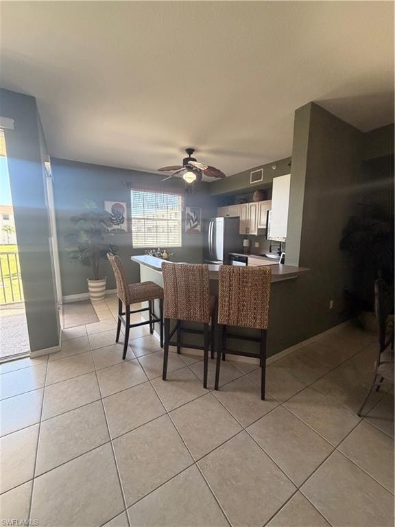 Open-concept kitchen featuring light-toned tile flooring, light wood-finish cabinetry, stainless steel appliances, and a breakfast bar with seating