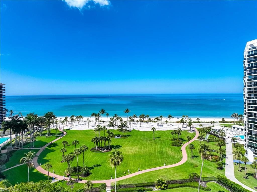 Expansive coastal landscape featuring white sand beaches, turquoise waters, a verdant lawn with mature palm trees, and paved pedestrian paths