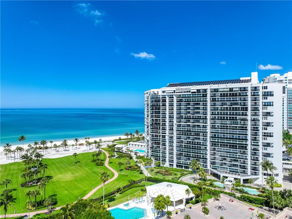 High-rise condominium building with multiple balconies, positioned alongside a white sand beach and expansive green space