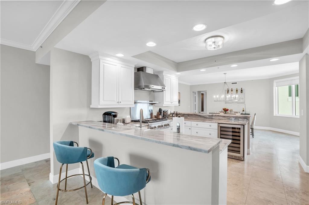 Kitchen with wine cooler, white cabinets, kitchen peninsula, wall chimney range hood, and light stone countertops