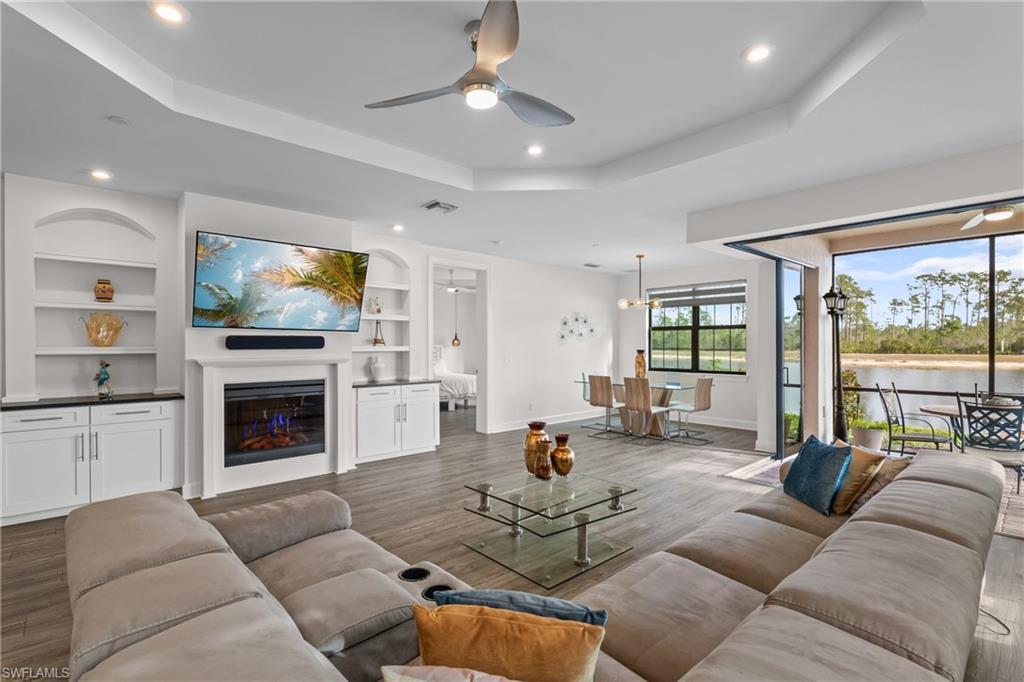 Living room with a ceiling fan, a glass covered fireplace, a raised ceiling, built in shelves, and dark wood-style floors