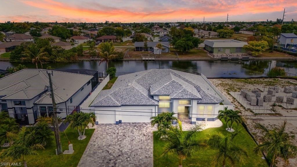 Waterfront property featuring a paver driveway, mature landscaping, multi-peak roofline, and an integrated two-car garage