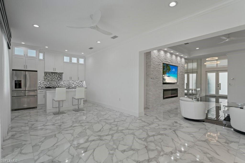 Open concept living space featuring large format porcelain tile flooring, a contemporary kitchen with white cabinetry and stainless steel appliances, a stone accent wall with an integrated fireplace and television mount, and recessed lighting throughout