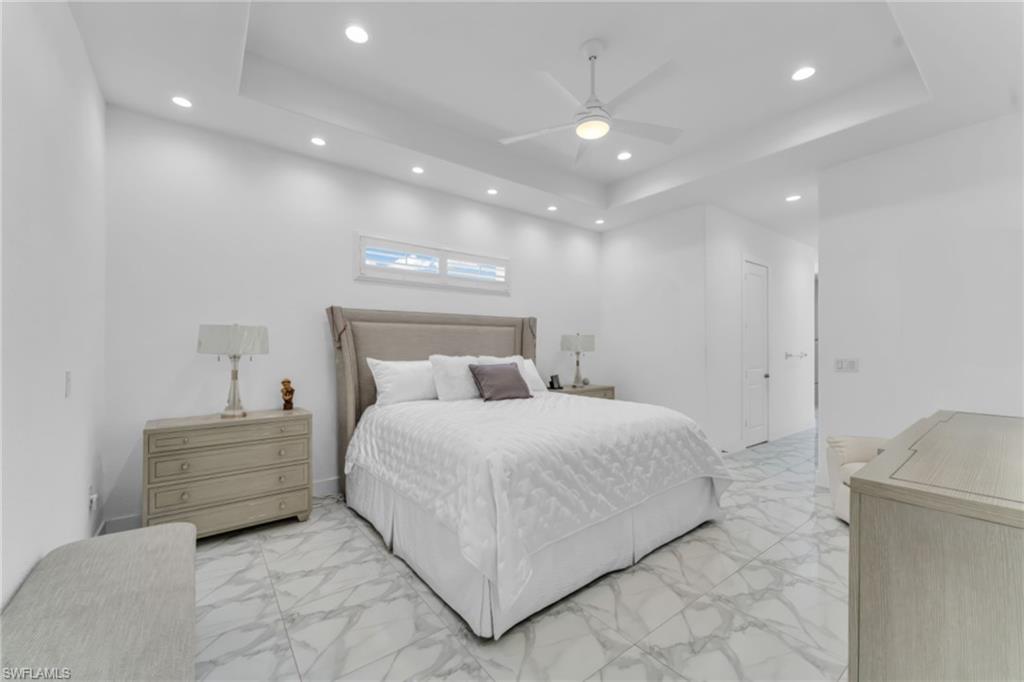 Bright room featuring white walls, a tray ceiling with recessed lighting and a ceiling fan, a headboard, a series of transom windows, and large format marble-look tile flooring